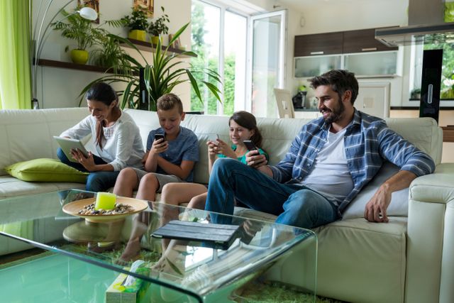 Family sitting on a white couch in a modern living room, each using different digital devices like a laptop, mobile phone, and tablet. The room is well-lit with natural light and decorated with green plants, creating a cozy and contemporary atmosphere. Ideal for use in articles or advertisements about family life, technology use at home, digital lifestyle, or modern home interiors.