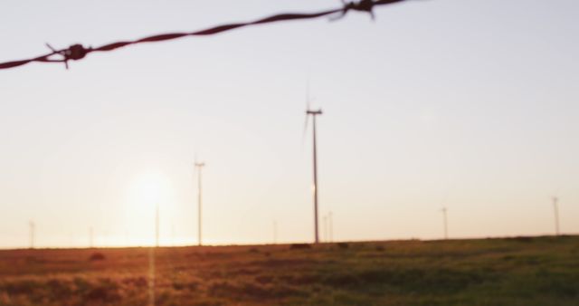 This image depicts multiple wind turbines standing prominently in a rural landscape at sunset, with a barbed wire fence in the foreground. Appropriate for illustrating themes related to renewable energy, environmental sustainability, and rural industry. Useful for websites, magazines, and promotional materials focusing on alternative energy sources and countryside living.