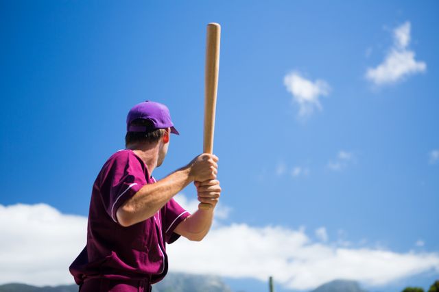 Baseball player in uniform holding bat, ready to swing, under clear blue sky on a sunny day. Ideal for sports-related content, outdoor activity promotions, athletic events, and competitive spirit themes.