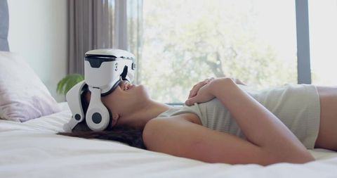 Woman lying wearing white vr headset and headphones, relaxing on bed by sunlit window