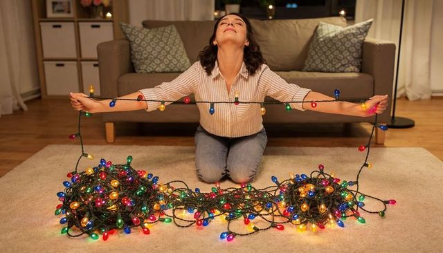 Woman kneeling untangling multicolored christmas string lights in cozy living room