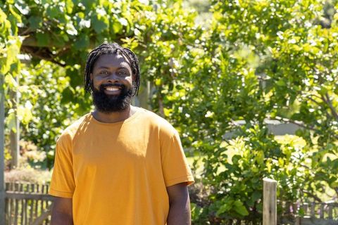 Sunlit african man smiling in backyard garden wearing orange t-shirt with dreadlocks and beard