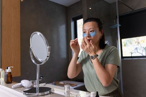 Mature asian woman applying blue under-eye hydrogel patches at bathroom vanity in daylight