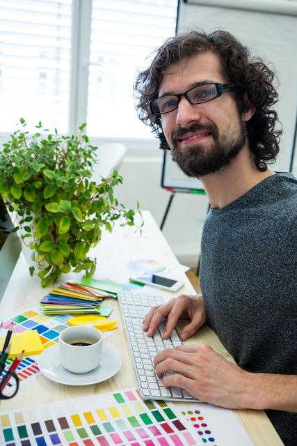 This image shows a male graphic designer working on a computer in a modern office. He is surrounded by color swatches, a cup of coffee, and a green plant, creating a vibrant and creative workspace. This image can be used for articles or advertisements related to graphic design, creative professions, modern work environments, or office decor.