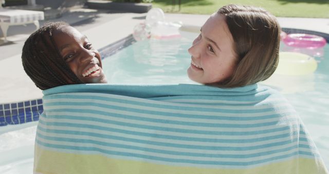 Two happy diverse teenage girl friends wrapped in towel sitting by sunny swimming pool. Friendship, weekend, free time, summer, vacations and togetherness, unaltered.