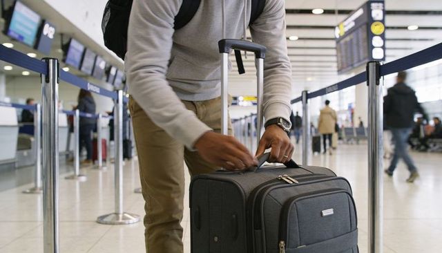 Traveler adjusting rolling suitcase handle in busy airport check-in queue preparing flight