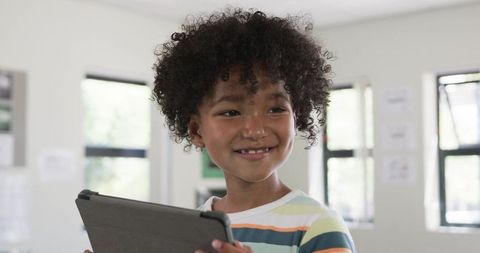 Curly-haired Schoolboy Holding Tablet and Smiling While Learning in Daylit Classroom