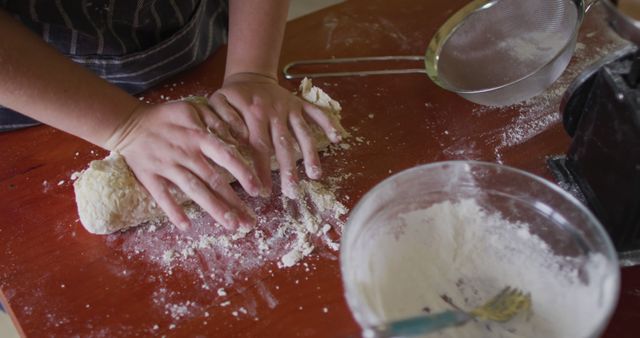 Close-up of person kneading dough on a wooden table sprinkled with flour. There is a glass bowl with flour, a fork, and a sieve in the frame, suggesting a bread or pastry-making process. Ideal for use in articles, blogs, or advertisements related to home baking, recipes, culinary classes, kitchen activities, and food preparation.