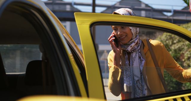 Front view of a young biracial woman wearing a hijab, talking on the smartphone and getting in a taxi in a city