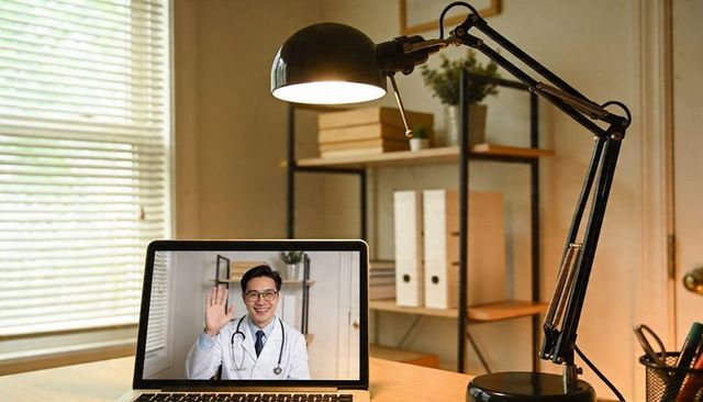 Waving doctor on laptop at home desk with task lamp for telemedicine consultation