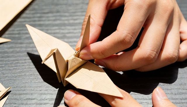 Adult hands folding brown origami crane on dark wooden table, closeup papercraft texture