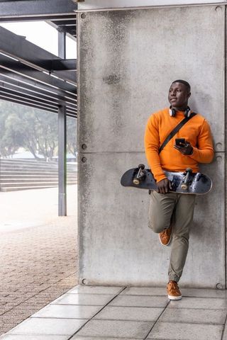 African american skateboarder leaning on concrete wall checking smartphone wearing orange sweater
