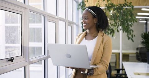 African american woman holding laptop near window smiling in open-plan office