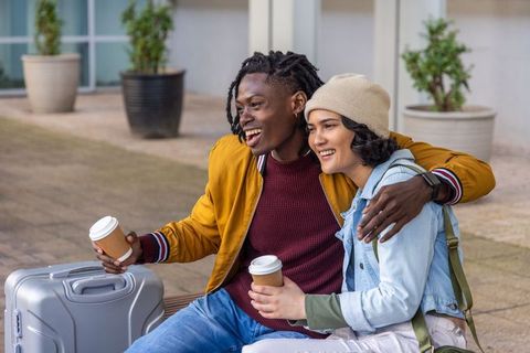 Laughing couple sharing takeaway coffee while traveling with suitcase and backpack