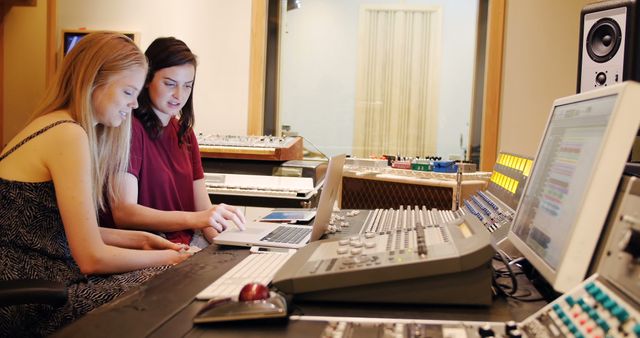 Two women are working together in a modern recording studio, analyzing tracks on a laptop and adjusting sound settings on a mixer. They are focused on their task, showcasing teamwork and creativity in a professional sound engineering environment. This image is ideal for illustrating concepts related to music production, collaboration, technology, and female professionals in audio engineering.