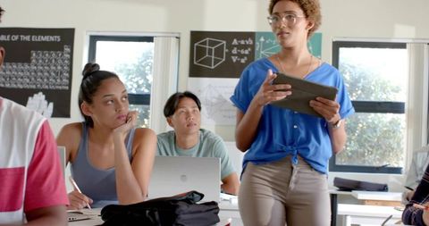 Asian woman presenting lesson with tablet while students taking notes in bright classroom