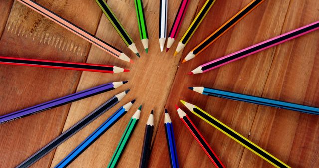 Colored pencils are arranged in a circular pattern on a wooden surface, creating a visually pleasing radial design. This arrangement showcases a variety of pencil colors, often used for artistic purposes or educational activities.