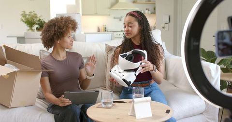 Two African American women unboxing VR headset on sofa filming live with ring light
