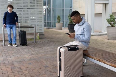Young black man checking phone while sitting with beige suitcase in modern transit plaza