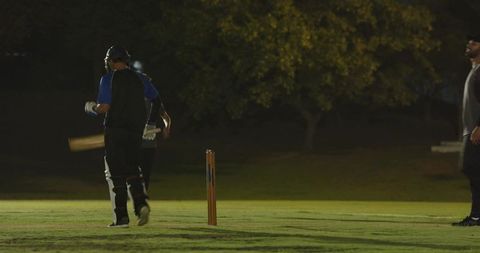 Night cricket batsman jogging between stumps under floodlights on grass pitch