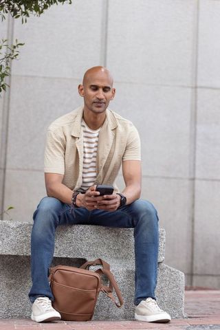 African american commuter sitting on urban bench holding smartphone and leather satchel