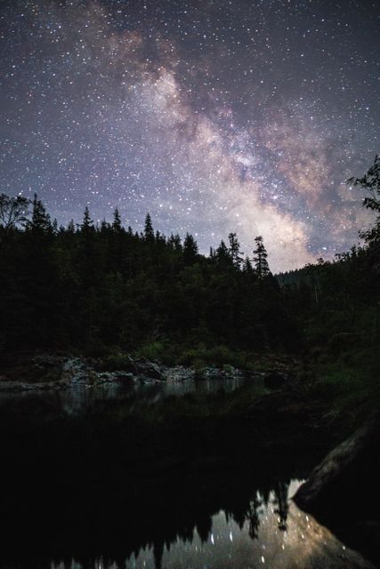 Milky Way arcs across night sky, reflected in calm lake surrounded by forest. Useful for astronomy blogs, nature photography articles, outdoor adventure marketing, and educational content featuring night sky and cosmos.