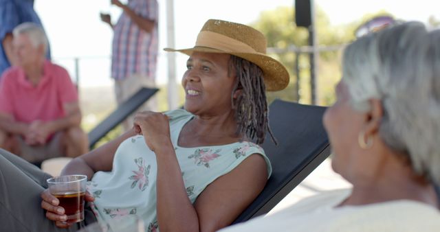 Senior African American women are relaxing on lounge chairs outdoors, smiling and enjoying a summer day with drinks in hand. Ideal for use in promoting senior lifestyle, leisure activities, vacations, and friendship among elderly people.
