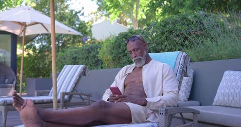 Senior african man relaxing poolside on chaise using smartphone, wicker furniture, garden