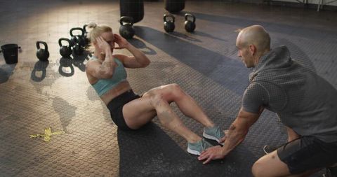 Female athlete performing sit-ups with personal trainer holding feet in functional gym
