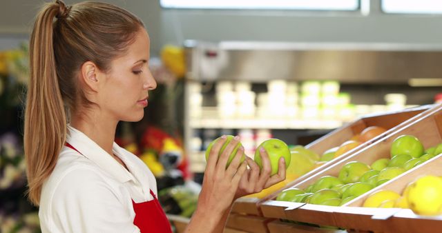 Image shows a female grocery store employee handling fresh green apples in a supermarket. She appears focused on examining the apples, highlighting the importance of quality control and fresh produce in retail. Ideal for use in articles or advertisements related to grocery shopping, fresh food, supermarkets, retail job roles, and customer service.