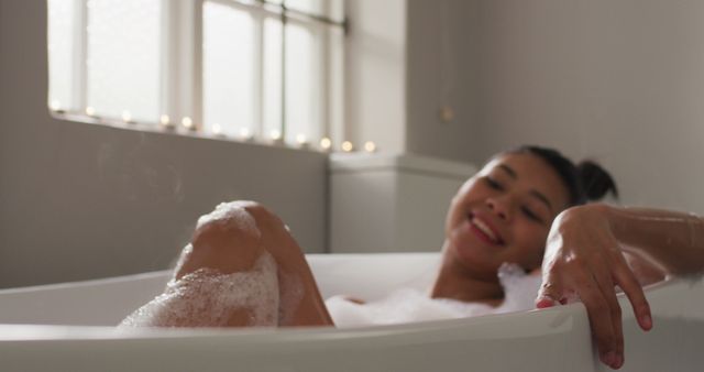 Woman lounging in bathtub filled with bubbles near sunlit window. Ideal for themes of self-care, wellness, relaxation, and spa retreats. Suitable for promotional materials for spas, bathing products, and relaxation therapies.