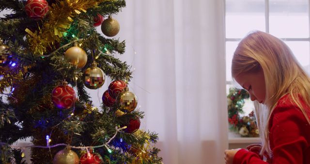Caucasian girl decorates a Christmas tree at home. She's immersed in the holiday spirit, adding ornaments with care.