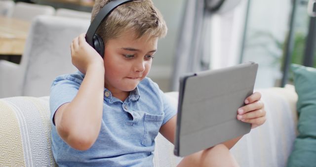 Young boy sitting indoors, using a tablet, and wearing headphones. His attention is focused on the tablet, suggesting he is either playing games, watching videos, or attending online learning sessions. This is suitable for illustrating concepts related to modern childhood, digital technology use, remote learning, home entertainment, and kids' leisure activities.