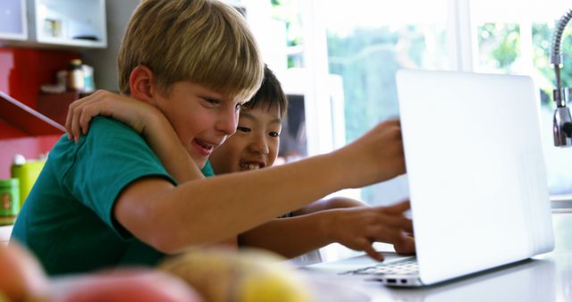 Two boys are engaging with a laptop in a brightly lit home kitchen. They are excitedly interacting with something on the screen, hugging and smiling. The focus of the image is on their enthusiastic expressions and the laptop, suggesting a fun and educational activity. This image is ideal for use in educational websites, homeschooling resources, or content related to children and technology.