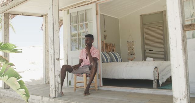 Thoughtful african american man sitting outside house on beach, copy space. Lifestyle, nature, relaxation, vacation, summer and leisure, unaltered.