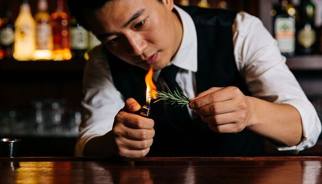 Bartender torching rosemary garnish for cocktail using lighter at intimate bar