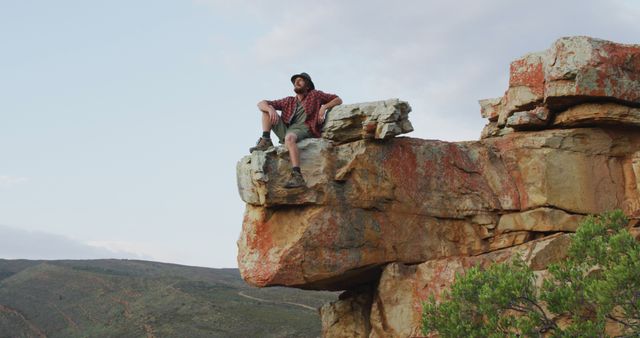 Man sitting calmly on large rock outcrop overlooking rugged landscape and hills. Perfect for illustrating themes of adventure, hiking, outdoor activities, freedom, and nature exploration. Suitable for use in outdoor adventure magazines, travel advertising, inspirational posters, and hiking gear promotions.