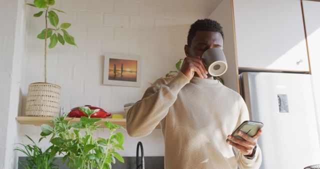 Happy african american man standing in kitchen, drinking coffee and using smartphone. Spending quality time at home alone.