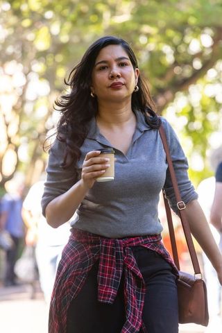 Woman walking with takeaway coffee, plaid shirt tied at waist, carrying brown shoulder bag