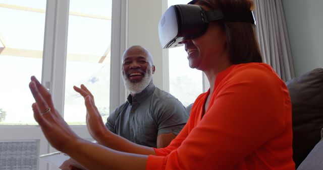 This image shows an older couple enjoying a virtual reality experience together at home, with one person using a VR headset and the other smiling. Useful for themes related to technology adoption among seniors, happiness, interactive experiences, and modern home lifestyle.