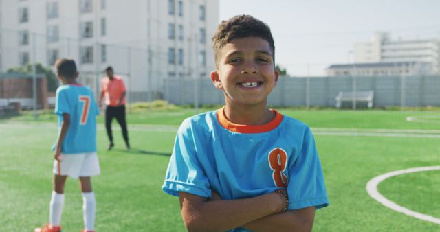 Boy smiling confidently on a soccer field in blue and orange jersey. Suitable for sports promotions, advertising athletic wear for children, and depicting team sports activities. Could be used in educational content on physical fitness and teamwork.