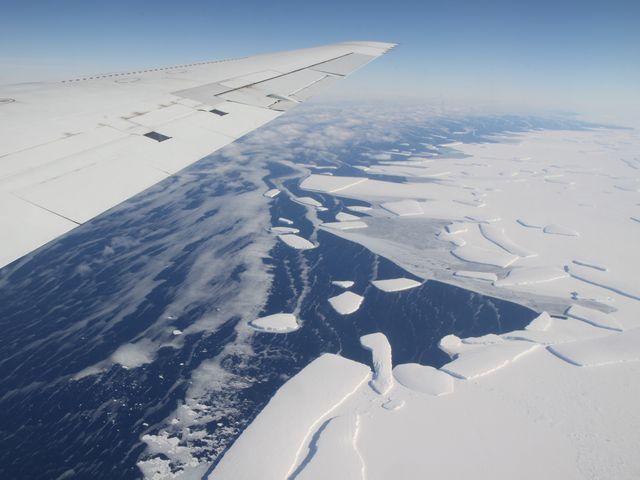 Aerial view from aircraft showing calving ice shelf in West Antarctica. White ice contrasts with dark ocean waters featuring floating ice chunks. Plane's wing in foreground indicates observation during flight, likely part of a research mission. Suitable for content on climate change, polar research, and NASA missions, as well as educational and scientific publications.