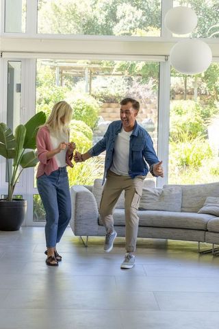 Middle-aged couple dancing joyfully in sunlit living room with garden view and modern sofa
