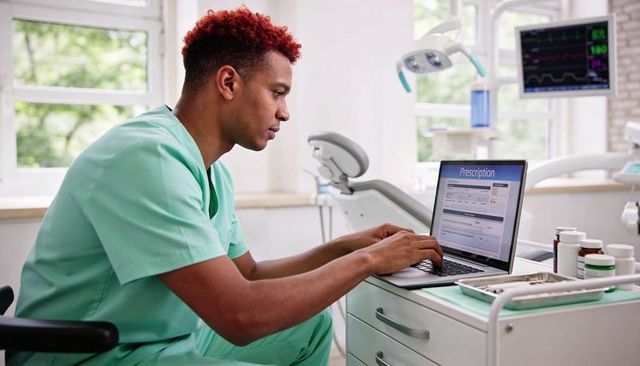 Clinician in green scrubs reviewing digital prescription on laptop in modern dental clinic
