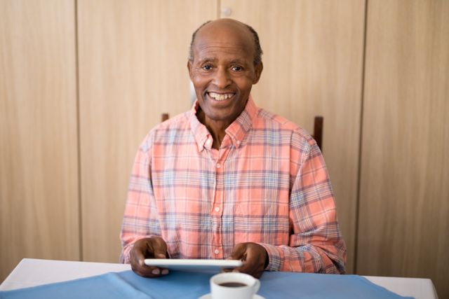 Senior man sitting at table in retirement home, smiling while using digital tablet. Coffee cup on table. Ideal for themes on senior lifestyle, technology use among elderly, independent living, and modern retirement.