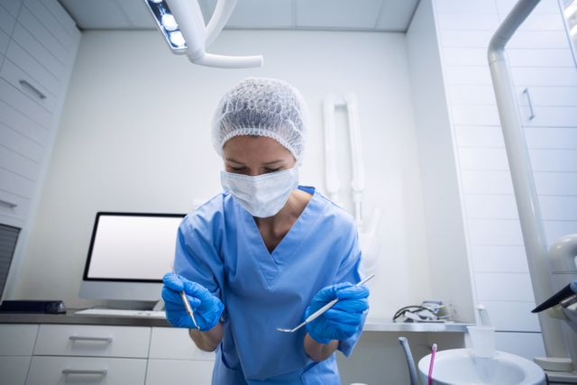 Dental assistant in blue scrubs holding dental tools, wearing surgical mask and gloves, in a modern dental clinic. This image can be used in healthcare contexts, dental care promotions, training materials for dental professionals, or medical equipment advertisements.