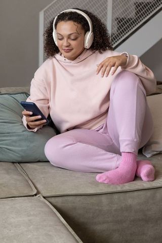 Relaxing at home: woman in pink loungewear listening to music and browsing phone on sofa
