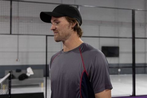 Male athlete standing by fenced indoor court wearing black cap and performance shirt
