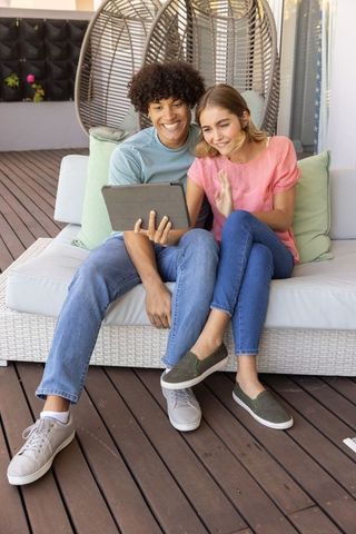 Smiling diverse young couple sharing tablet and waving while relaxing on rattan sofa