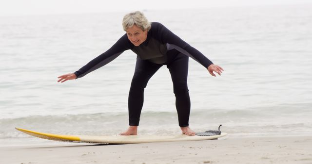 Senior woman dressed in a wetsuit practicing balancing on a surfboard on sandy beach. Perfect for illustrations of active elderly lifestyle, fitness for older adults, and outdoor hobbies. Suitable for advertisements promoting active living and health products for seniors.